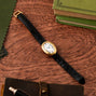 The Cartier Baignoire gold watch with a black leather strap rests on a wooden desk next to green books and a brown notebook.