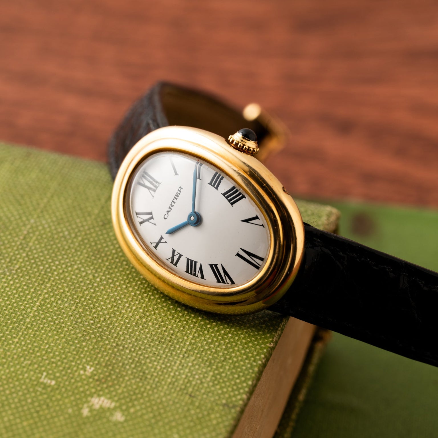 Cartier Baignoire gold wristwatch with black leather strap placed on a green textured book cover.