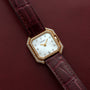 The Cartier Ceinture Diamond watch features diamond accents and a burgundy leather strap, showcased on a matching burgundy surface.