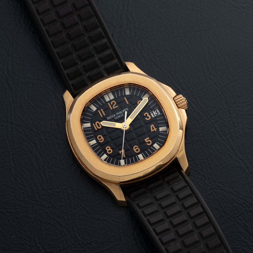 Patek Philippe Aquanaut watch with a black strap and black dial, displayed on a dark textured surface.