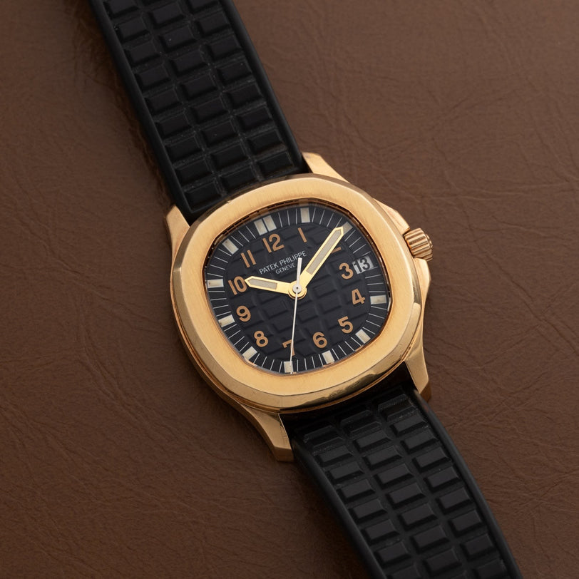 Patek Philippe Aquanaut wristwatch with a black strap and black dial, displayed on a brown leather surface.