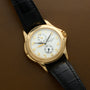 The Patek Philippe Calatrava Travel Time wristwatch with a black leather strap rests on a brown surface, featuring Arabic numerals and subdials.