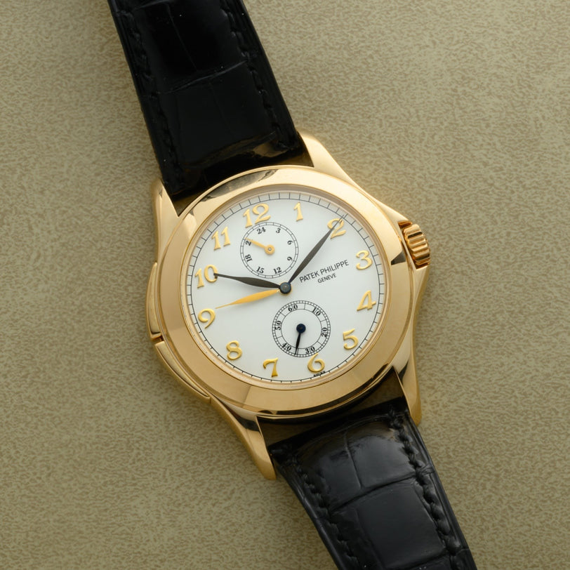 A Patek Philippe Calatrava Travel Time gold wristwatch with a white dial and black leather strap displayed on a beige surface.