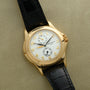 A Patek Philippe Calatrava Travel Time gold wristwatch with a white dial and black leather strap displayed on a beige surface.