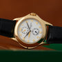 The Patek Philippe Calatrava Travel Time features a gold case, white dial, black leather strap, and Arabic numerals, set against a brown background.