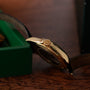 The Patek Philippe Calatrava Travel Time gold wristwatch with a black leather strap rests on a wooden surface beside a green box.