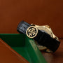 A Patek Philippe Calatrava Travel Time watch with a black leather strap and gold emblem rests on a green and brown surface.