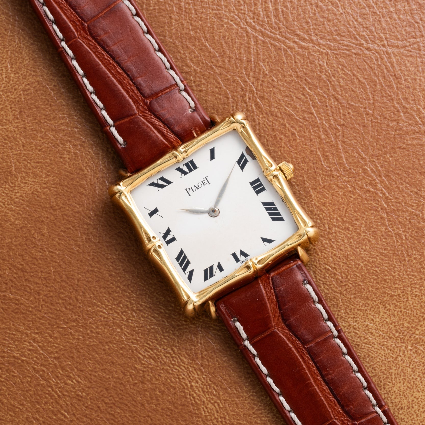 The Piaget Bamboo Dress Watch features a gold square case, Roman numerals, and a brown leather strap, displayed on a tan leather surface.