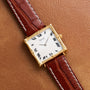 The Piaget Bamboo Dress Watch features a gold square case, Roman numerals, and a brown leather strap, displayed on a tan leather surface.