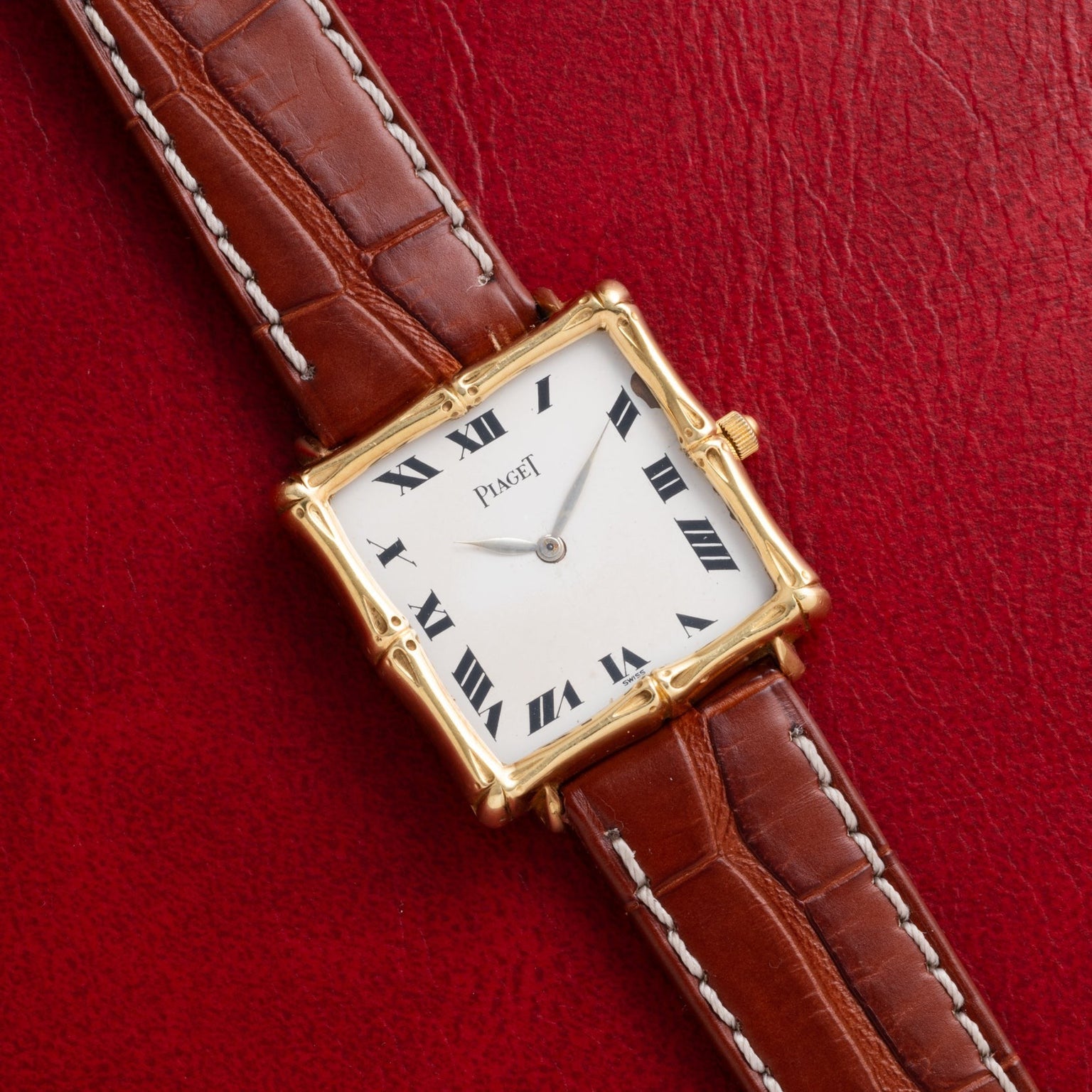 A Piaget Bamboo Dress Watch featuring a gold square case and brown leather strap, displayed on red textured leather.