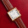 A Piaget Bamboo Dress Watch featuring a gold square case and brown leather strap, displayed on red textured leather.