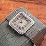 The Cartier Santos Platinum, a vintage wristwatch with Roman numerals, is displayed on a textured gray cushion atop a wooden surface.