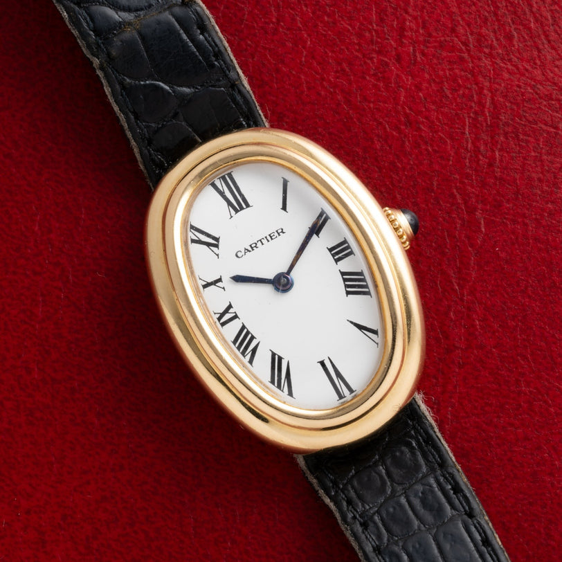The Cartier Baignoire watch features a gold oval case, Roman numerals, and a black leather strap, displayed on a red textured surface.