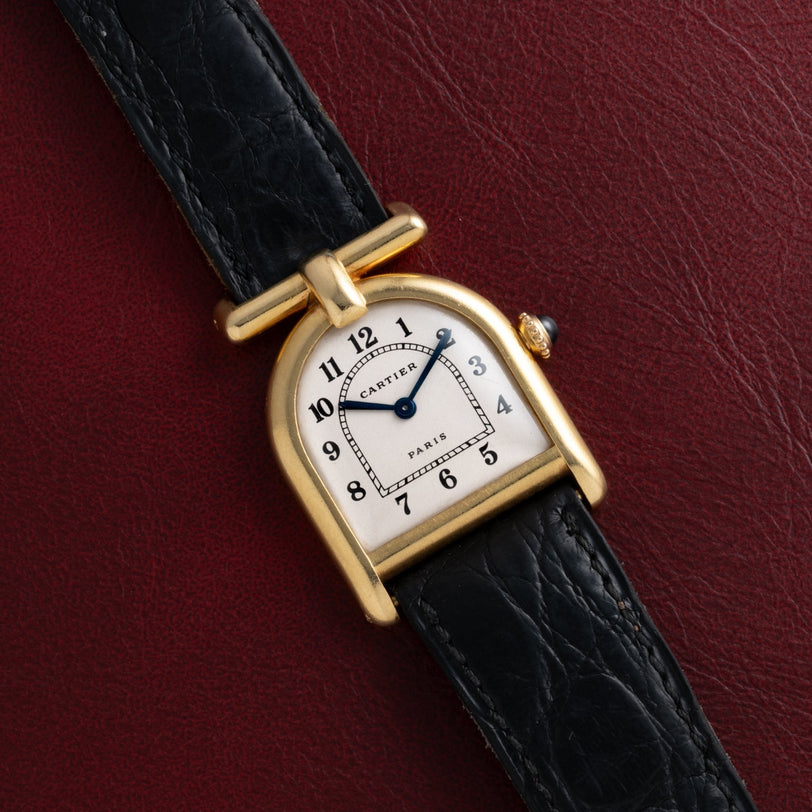 Cartier Calandre gold wristwatch with a black leather band displayed on a dark red textured surface.