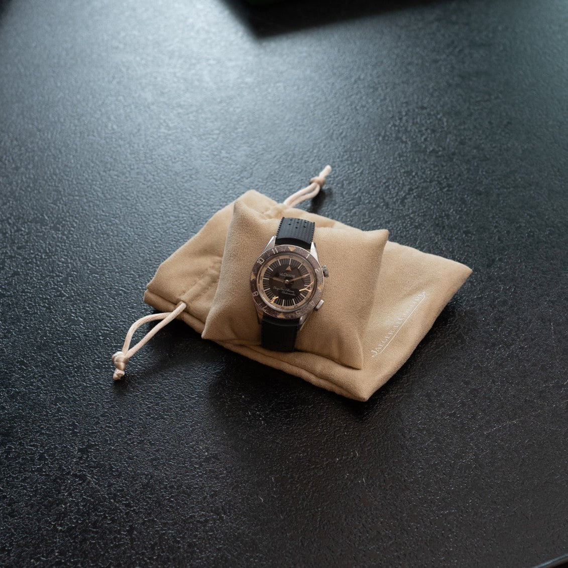 The Jaeger-LeCoultre Deep Sea Alarm U.S. Edition wristwatch with a black strap is displayed on a beige drawstring pouch set atop a dark textured surface.