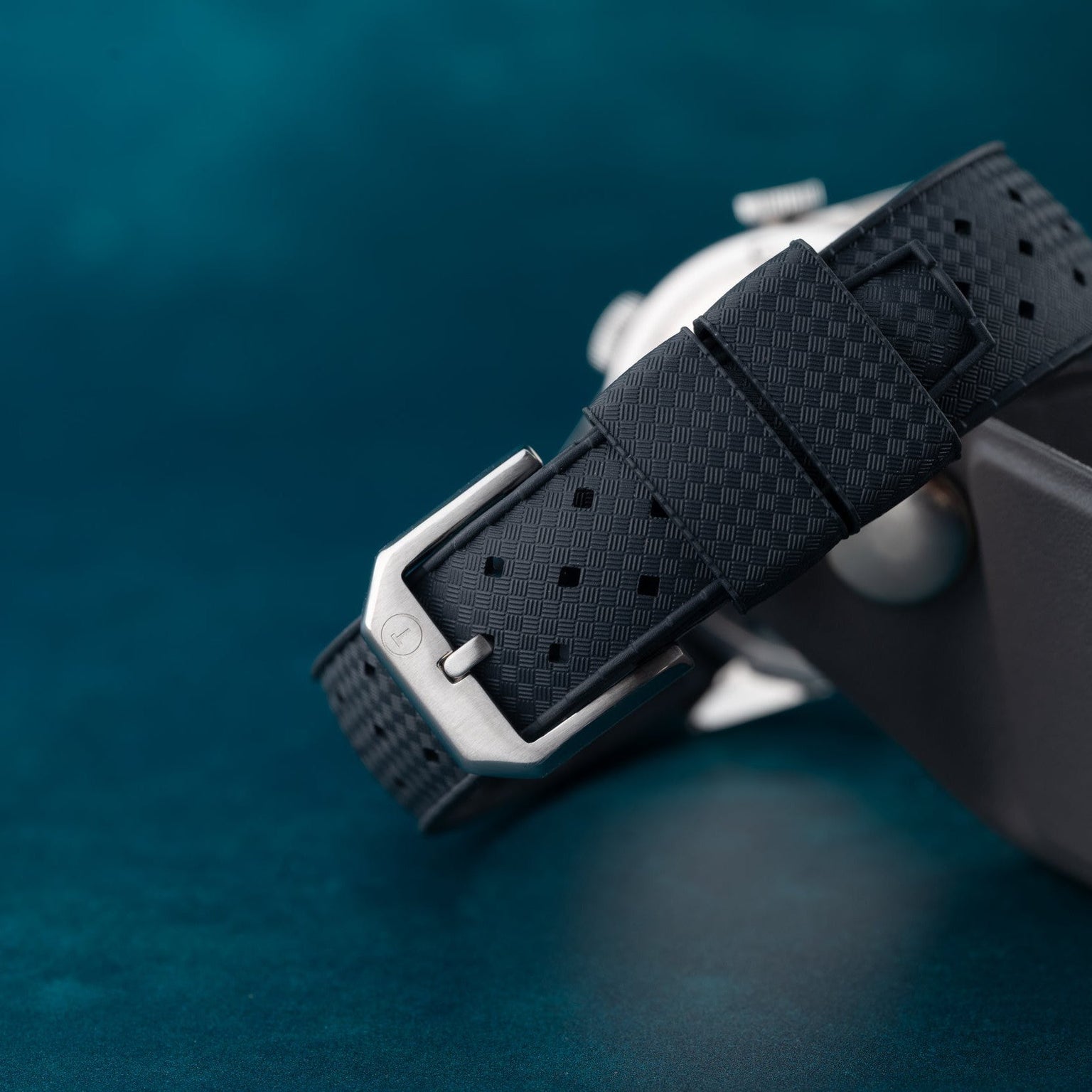 Close-up of the Jaeger-LeCoultre Deep Sea Alarm U.S. Editions black textured strap with a silver buckle, set against a teal background.