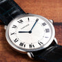 A Cartier Vendome Ronde Louis wristwatch with a white dial, Roman numerals, and blue hands on a leather strap, displayed on wood.