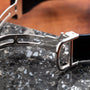 Close-up of a silver Cartier Vendome Ronde Louis watch clasp on a black band, resting on a speckled stone surface.