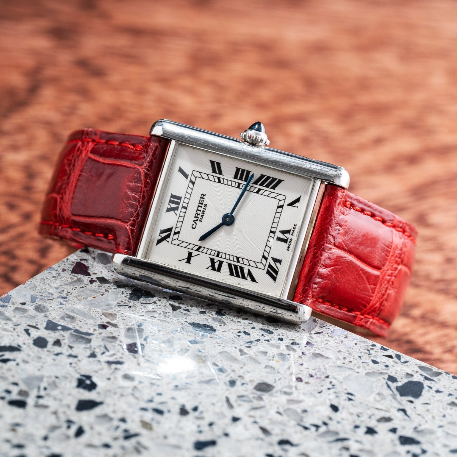 A Cartier Tank Louis watch with a red leather strap and Roman numerals rests on a terrazzo surface.