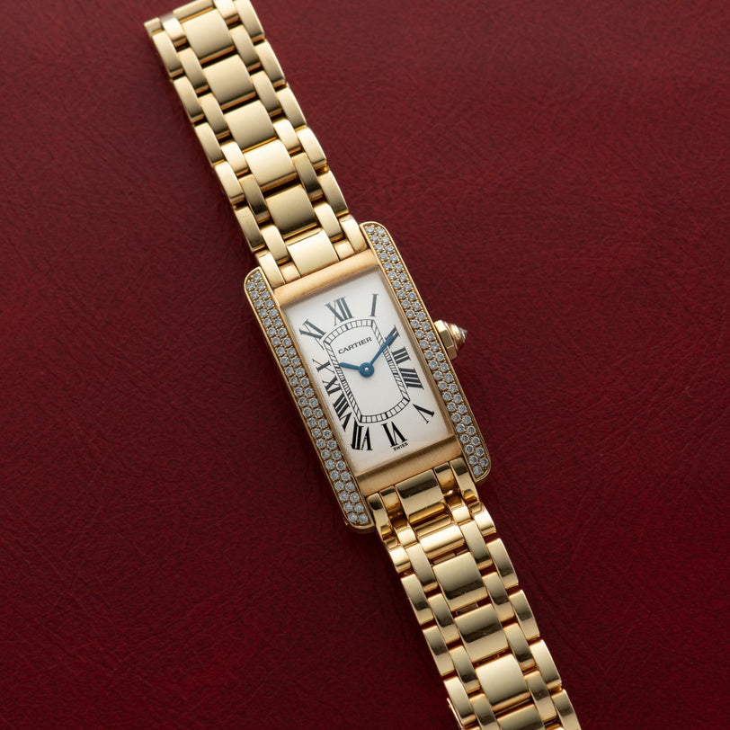 The Cartier Tank Americaine Diamond watch features a rectangular face with diamond accents, showcased on a red textured surface.