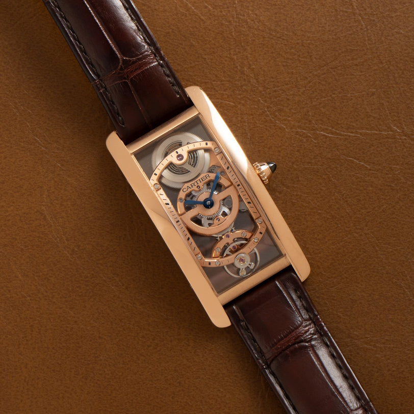 The Cartier Tank Cintreé Skeleton is a rectangular gold watch with a skeleton dial and brown leather strap, displayed on a brown leather surface.