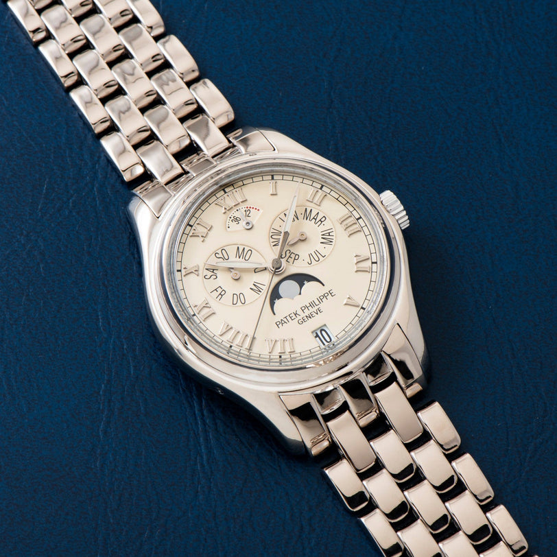 A Patek Philippe Annual Calendar silver wristwatch with a metal band rests on a dark blue textured surface.