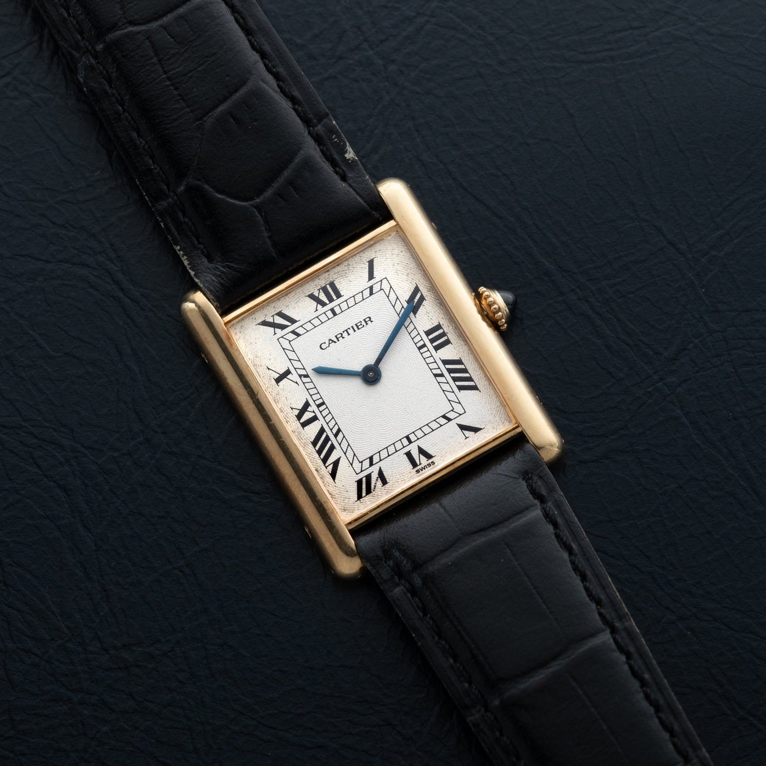 The Cartier Tank Louis Mechaniqúe features a gold case, black leather strap, and Roman numerals on a textured black dial.