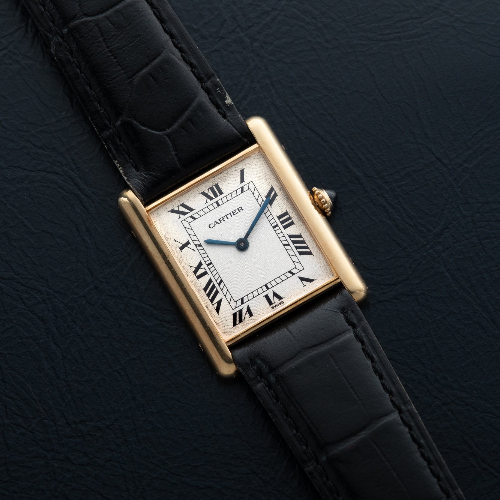 The Cartier Tank Louis Mechaniqúe features a gold case, black leather strap, and Roman numerals on a textured black dial.