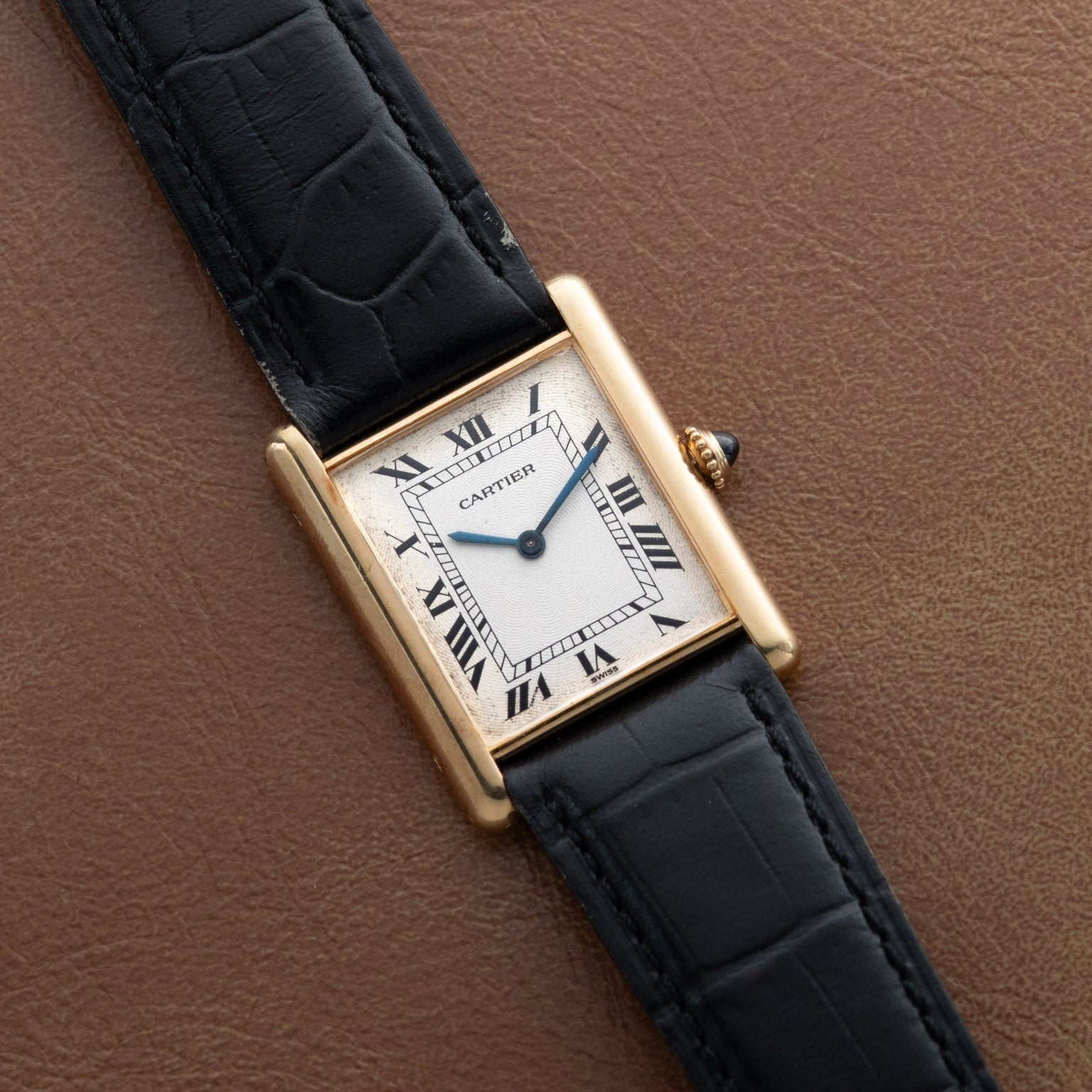 A Cartier Tank Louis Mechaniqúe gold watch with a black leather strap rests on a brown leather surface.