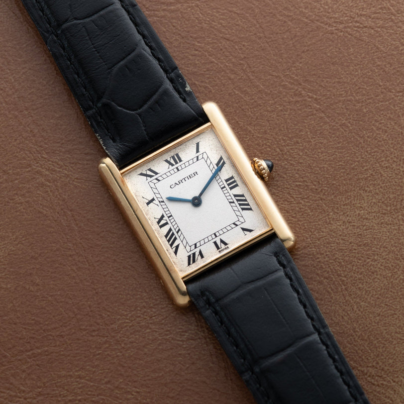 A Cartier Tank Louis Mechaniqúe gold watch with a black leather strap rests on a brown leather surface.