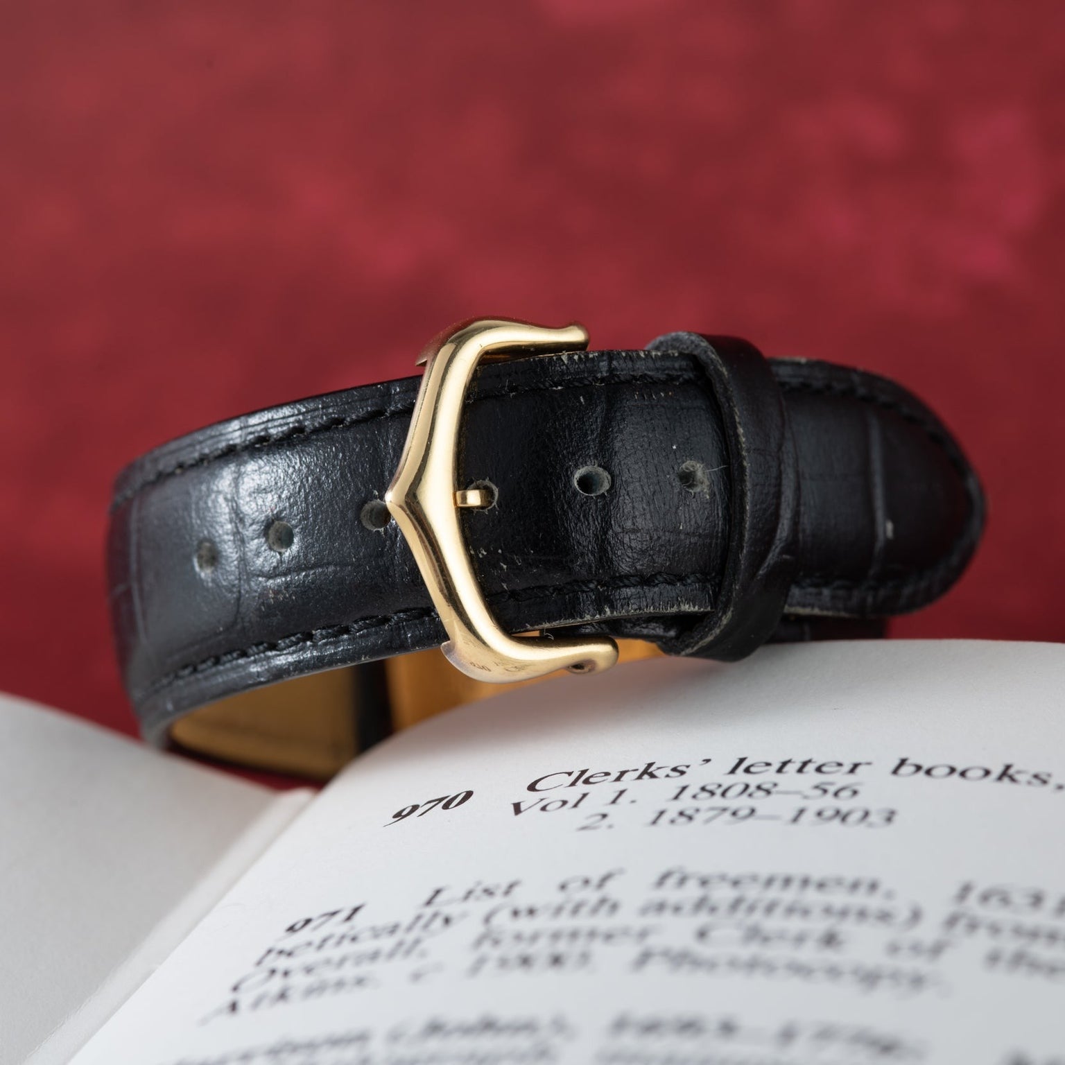 The Cartier Tank Louis Mechaniqúe black leather strap with a gold buckle rests on an open book against a red backdrop.