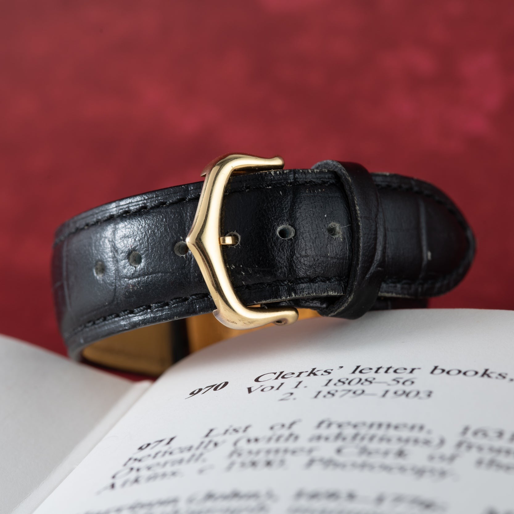 The Cartier Tank Louis Mechaniqúe black leather strap with a gold buckle rests on an open book against a red backdrop.