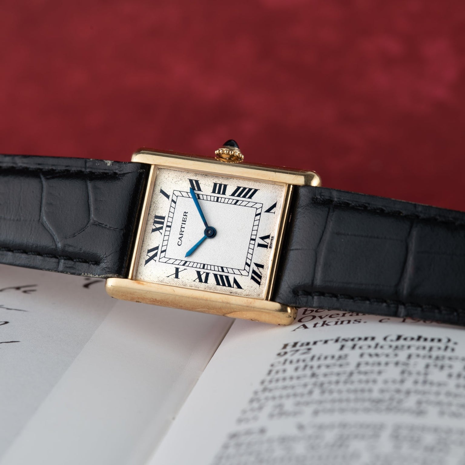 A Cartier Tank Louis Mechaniqúe gold watch with a black leather strap rests on an open book with text, set against a red background.