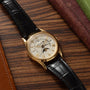 The Patek Philippe Retrograde Perpetual Calendar wristwatch, featuring a black leather strap, is displayed on a wooden surface beside brown and green books.
