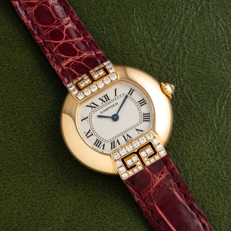 Cartier Ellipse watch with Roman numerals, diamond accents, and a red leather strap shown on a green textured surface.