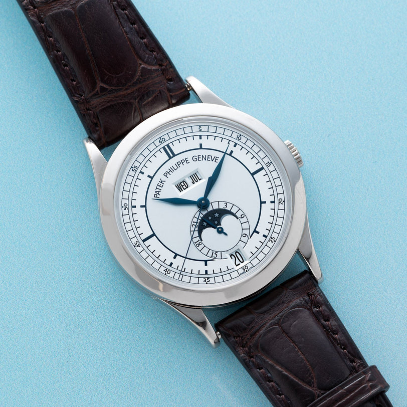 Patek Philippe Annual Calendar watch with white face, moonphase indicator, and dark brown leather strap displayed on a light blue background.