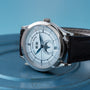The Patek Philippe Annual Calendar luxury wristwatch features a white dial, black leather strap, and calendar display, shown on a blue surface.