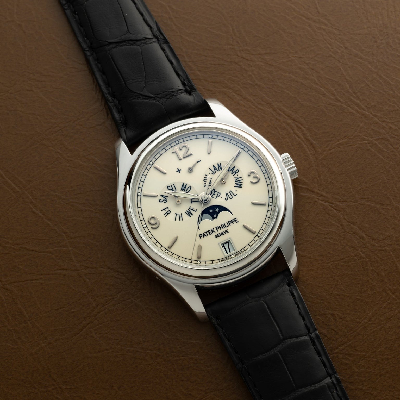 The Patek Philippe Annual Calendar Moonphase luxury watch features a black leather strap and displays moonphase and calendar functions, shown here on a brown surface.