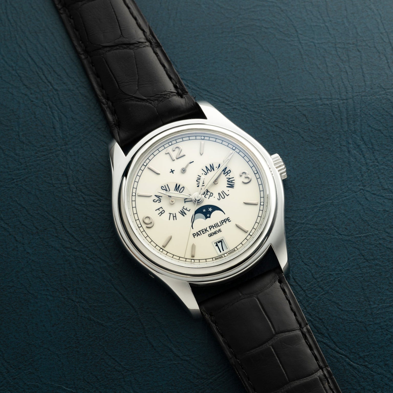 The Patek Philippe Annual Calendar Moonphase watch with a black leather band is displayed on a textured dark green surface.