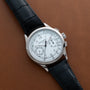 A Patek Philippe Chronograph wristwatch with a white dial and black leather strap displayed on a brown surface.