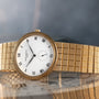 The Patek Philippe Calatrava, featuring a white face with Roman numerals and a patterned gold band, rests on a reflective surface.