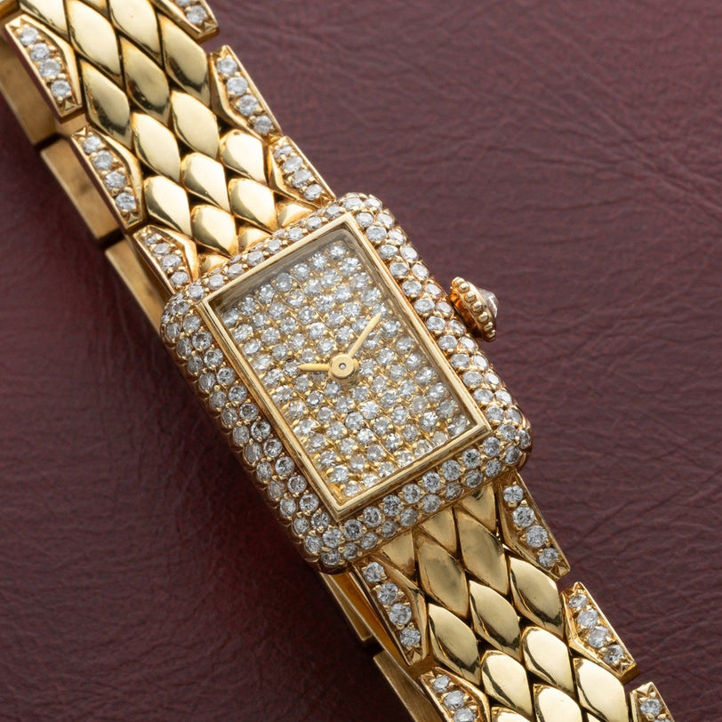 The Cartier Tank Mini Diamond wristwatch features a rectangular, diamond-studded face and band, elegantly displayed on a burgundy surface.