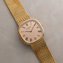 A Patek Philippe Ellipse Jumbo gold wristwatch with Roman numerals and a mesh band rests on a light textured surface.