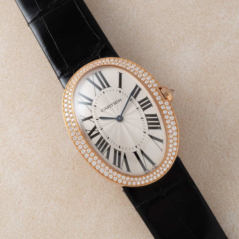 A Cartier Baignoire Jumbo watch featuring an oval case, diamond bezel, and black leather strap displayed on a beige surface.