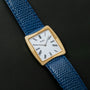 A gold Cartier Protocole watch with a white face, Roman numerals, and a blue textured leather strap on a black background.