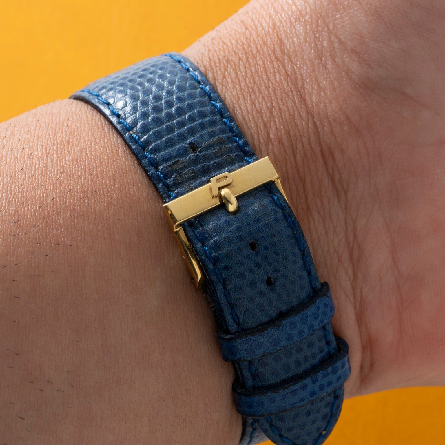 Close-up of a wrist wearing the Cartier Protocole watch with a blue textured strap and gold clasp, set against a yellow background.