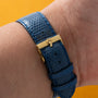 Close-up of a wrist wearing the Cartier Protocole watch with a blue textured strap and gold clasp, set against a yellow background.