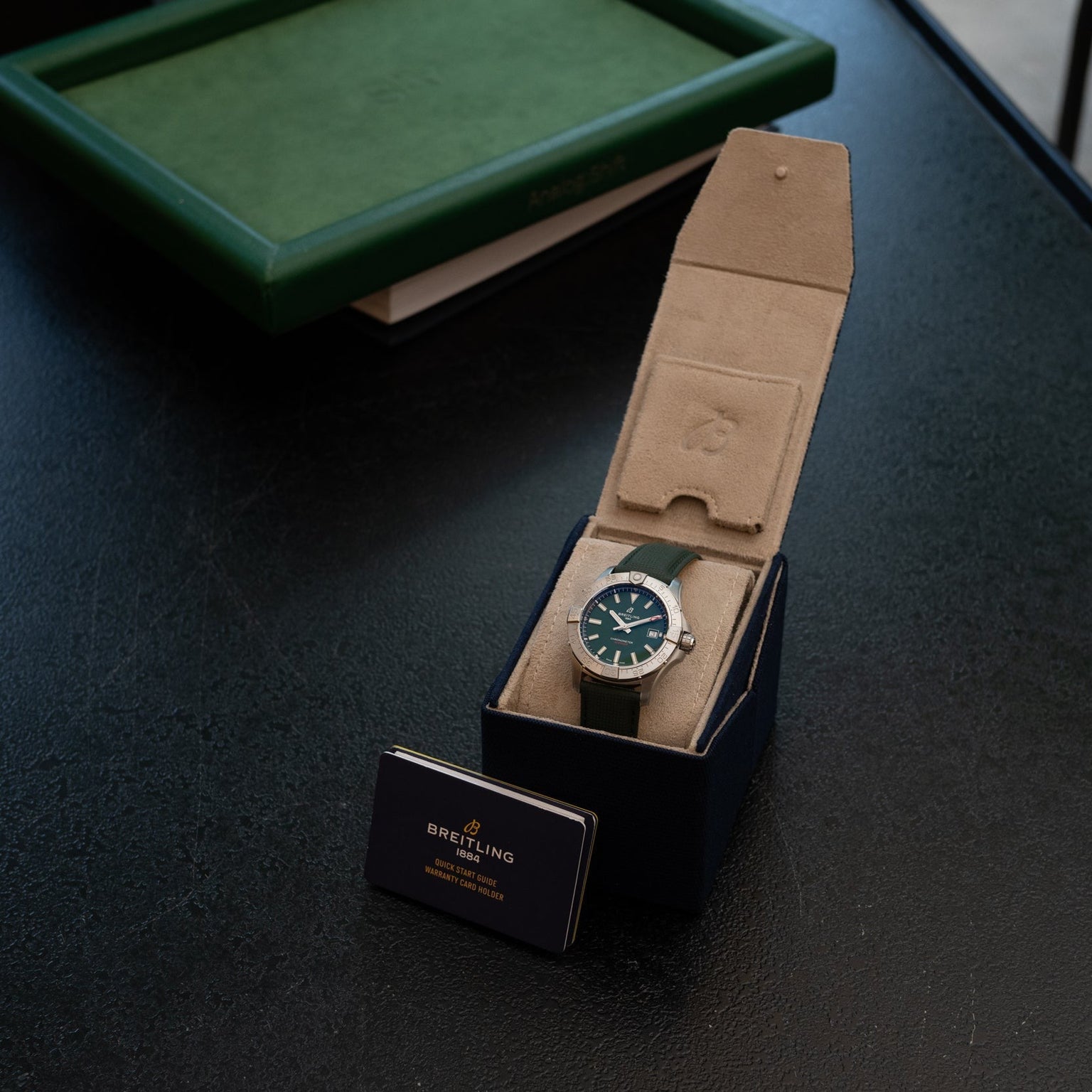 A Breitling Avenger Automatic 42 watch with a green dial in a tan case is displayed beside a Breitling card on a dark table.