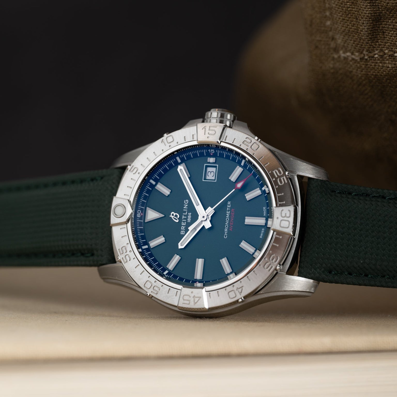 A Breitling Avenger Automatic 42 with a silver case, blue dial, and green strap rests on a light surface.