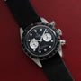 A TUDOR Black Bay Chronograph watch with a black strap displayed on a red textured surface.